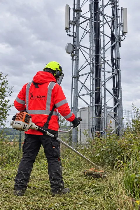 Technicien ALTICOM débroussaillant au pied d'un pylône télécom